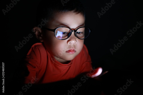 The boy uses a smartphone at night. His face, framed by glasses, stares at the cell phone screen. Laptop technology. A brightly lit screen. Light screen
