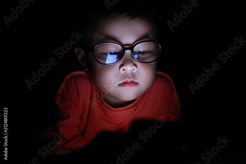 The boy uses a smartphone at night. His face, framed by glasses, stares at the cell phone screen. Laptop technology. A brightly lit screen. Light screen