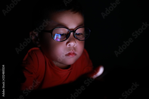 The boy uses a smartphone at night. His face, framed by glasses, stares at the cell phone screen. Laptop technology. A brightly lit screen. Light screen