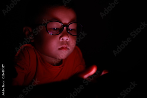 The boy uses a smartphone at night. His face, framed by glasses, stares at the cell phone screen. Laptop technology. A brightly lit screen. Light screen