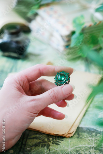 A woman is holding a diamond brooch with a large emerald at its center. A family heirloom set against a backdrop of love letters, a precious jewel.
