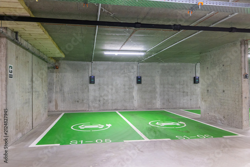 Electric cars parking lot. Modern underground concrete parking garage featuring bright green spaces marked with white electric vehicle charging symbols and wall-mounted EV chargers.