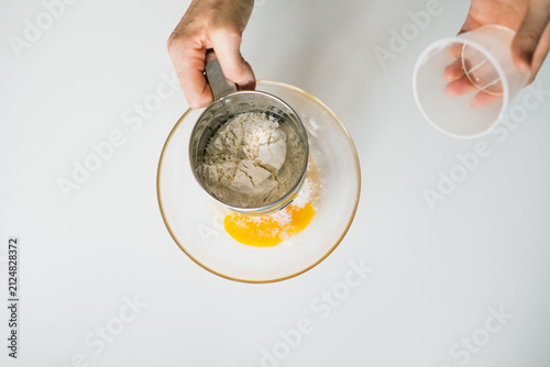 Fotografie Hand holding flour sifter over egg yolk in glass bowl for baking