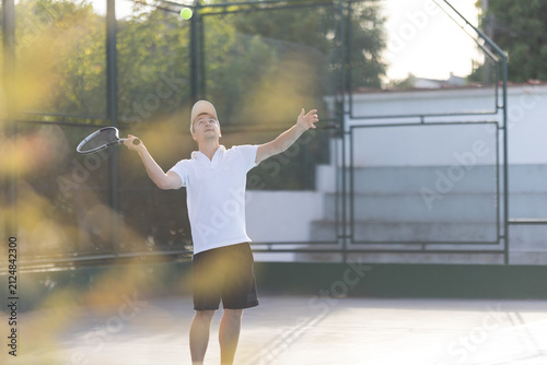 Man serves a tennis ball on a court during late afternoon practice session in a park