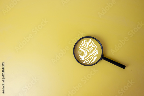 Cyber security and biometric authentication concept. A magnifying glass focusing on a white fingerprint icon graphic on a solid yellow background.