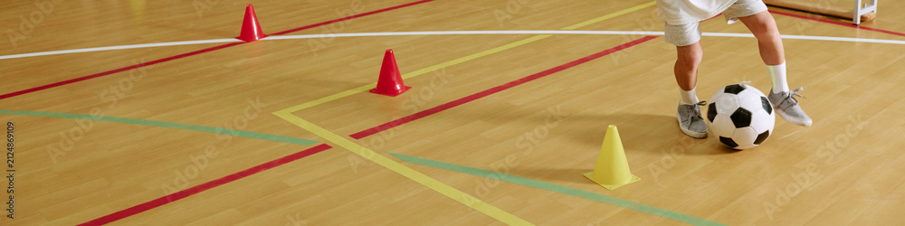 Fototapeta premium Header cropped shot of child practicing soccer dribbling around cones on indoor gym floor. Image suitable for youth sports programs, physical education, and kids fitness marketing