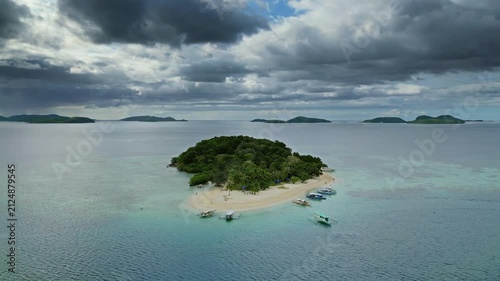 Aerial around view of small tropical island with clear sea waters and lush greenery with distant islands, dark moody sky, Philippines, 4k