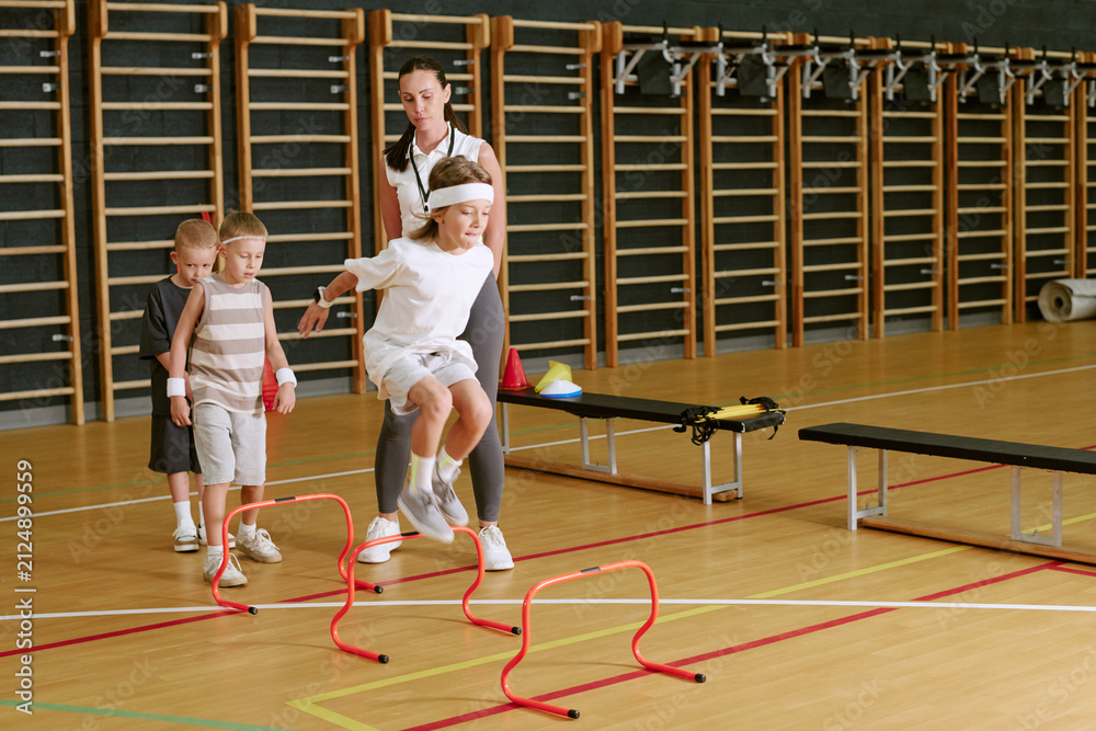 Fototapeta premium Caucasian female trainer supervising children jumping over hurdles in school gym. Image suitable for youth sports programs, physical education marketing, and healthy lifestyle campaigns