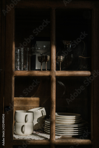 Vintage wooden cabinet displaying ceramic plates, cups and antique glassware in warm natural light. Cozy interior still life with timeless home aesthetics.