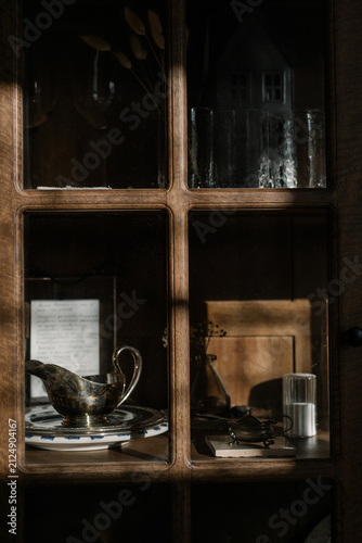 Vintage wooden cabinet displaying ceramic plates, cups and antique glassware in warm natural light. Cozy interior still life with timeless home aesthetics.