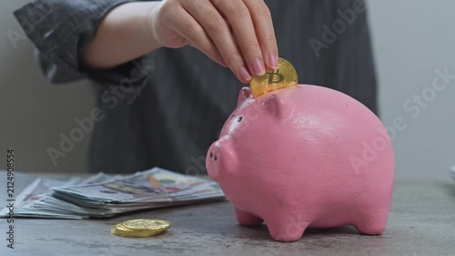 Woman puts gold bitcoin into pink piggy bank on desk next to cash and coins