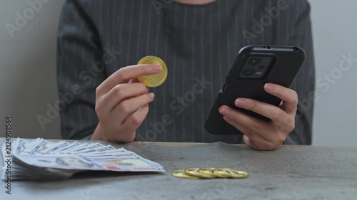 Woman holds gold bitcoin while checking smartphone with stacks of cash and crypto coins on desk