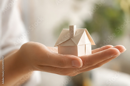 A close-up shot of a human hand delicately holding a miniature paper model house, showcasing a simple architectural design with a triangular roof and windows, creating a cozy and inviting scene.