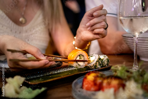 Hand holds baked sushi roll with chopsticks above shared table. Japanese sushi concept for family dining and Asian food culture.