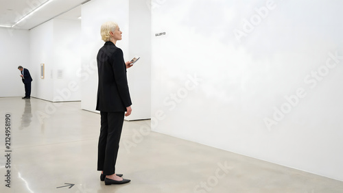 A short-haired woman in a black suit standing in a white gallery, holding a small and looking at a blank wall in a bright room space
