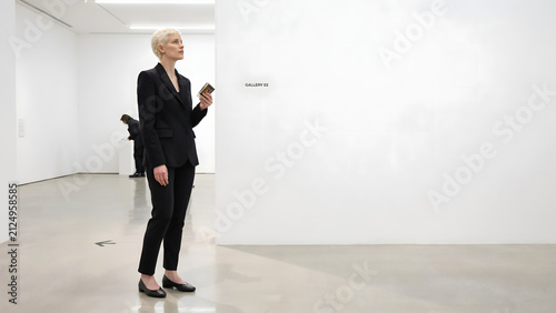 A short-haired woman in a black suit standing in a white gallery, holding a small and looking at a blank wall in a bright room space