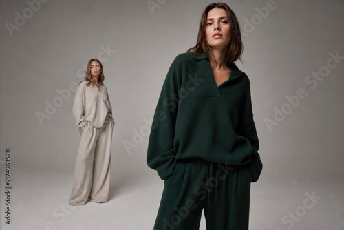Two women in modern oversized loungewear, relaxed pose against neutral studio backdrop, minimalist fashion photography with soft natural lighting, casual chic style