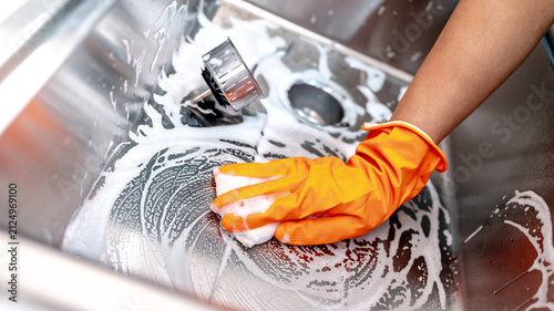 Obraz na płótnie Housekeeper hand wearing orange rubber glove scrubbing on kitchen sink drainer with soap suds spreading around