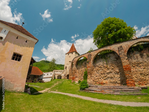 Photography Low angle photograph of the defensive stone walls, arches, and medieval towers of Biertan Fortified Church under a bright summer sky in Transylvania, Romania
