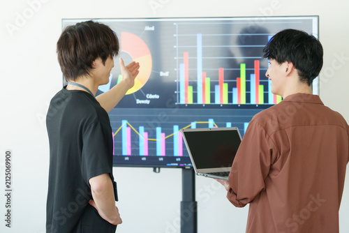 オフィスでディスプレイを前に会話する若い日本人男性2人の背中