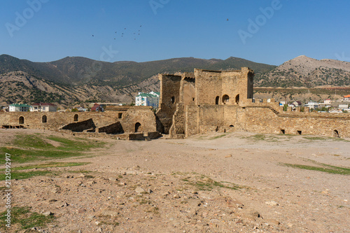 Ancient stone Genoese Fortress nestled in a valley, surrounded by rolling hills and modern buildings. Its crumbling walls and towers showcase historical architecture. Sudak. Crimea