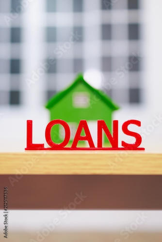 Картината върху платно Red loans letters in front of a green house model with blurred building background, representing mortgage, home financing and property lending