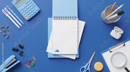 Flat lay of an open blank spiral notebook surrounded by office supplies including pens, calculator, scissors, and paper clips on a blue background.