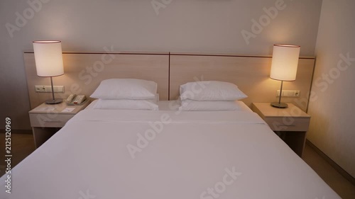 Clean hotel room with crisp white bedding, wooden headboard, matching nightstands. Warm lamp light creates cozy ambiance