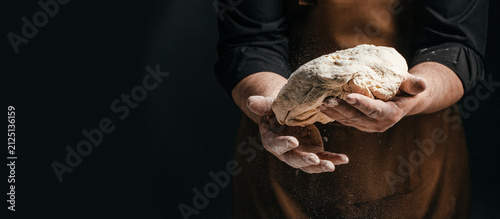 Fotografía Male baker in brown apron holds kneaded dough with flour dusting in hands, dark
