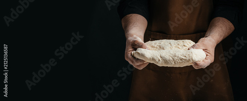 Cuadro en lienzo Male baker in brown apron holds dough ball with flour on hands, dark background