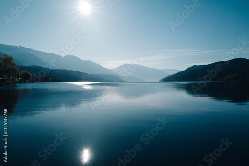Fotografía Sun blazing over a calm mountain lake with hazy blue ridges receding into the di