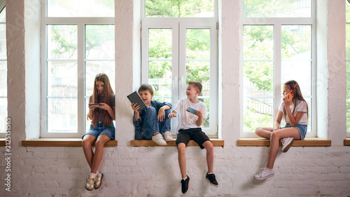 Quadro su tela Four school-age children using personal devices while sitting apart by window
