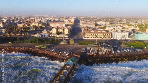 Aerial view of Swakopmund, Namibia, shows the pier, rocky breakwater, palm promenade, German colonial buildings, cafes, and moving people and cars in warm golden hour light.