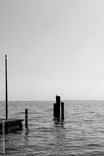 In Italy, mooring posts and a small pier form a black and white minimalist scene on Lake Garda, surrounded by calm water and open sky.