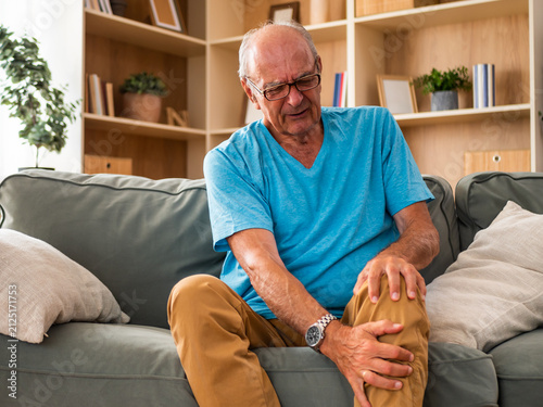 Worried senior man sitting on sofa in living room, massaging his painful knee due to arthritis and joint problems. Senior healthcare and aging lifestyle concept.