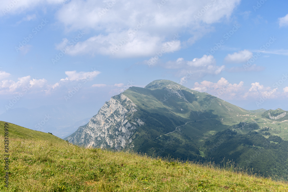 Fototapeta premium In Italy, a green mountain ridge with a pale rocky face rises across Monte Baldo, surrounded by meadow, forest patches and scattered summer clouds.