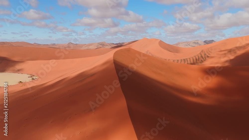 Aerial camera sweeps along a sharp dune ridge in Sossusvlei, Namibia, showing ripple patterns, a pale clay pan, and rocky ridges under bright sky and warm sand.