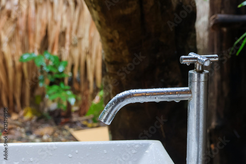 Konstfotografi Shiny metal faucet with water droplets over white basin in outdoor garden settin