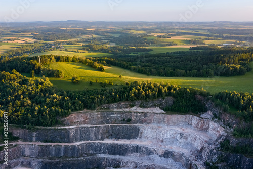 Aerial view of a large open pit stone quarry mine with terraced rock layers surrounded by green landscape during the sunset
