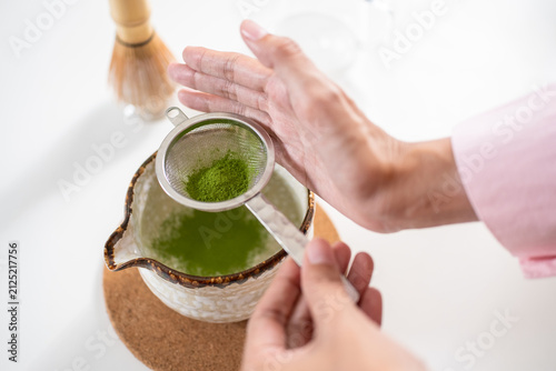 Картината върху платно High-angle close-up of hands tapping a metal strainer to sift organic matcha green tea powder into a ceramic bowl