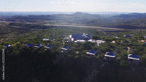 Aerial view of a Namibia cliff top lodge, blue roof chalets, pool, and ridge. The camera sweeps across the escarpment, roads, reservoir, and rolling hills.