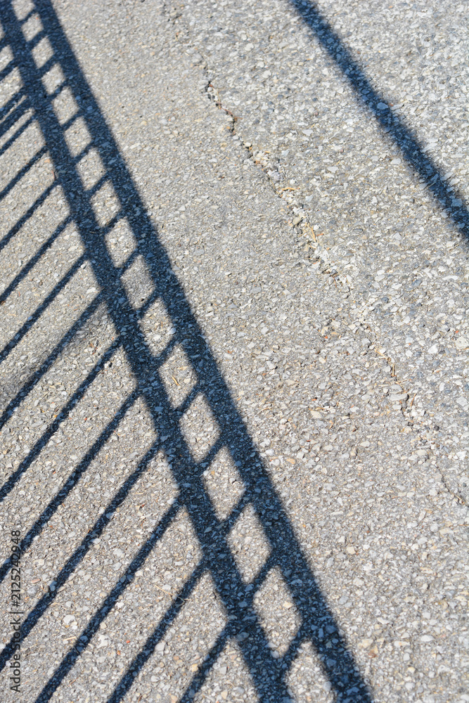 Fototapeta premium A high angle shot of a geometric shadow cast by a metal railing fence on a rough asphalt surface during a sunny day.