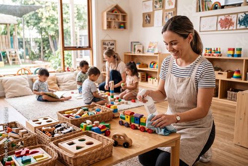 An educator disinfects wooden toys on a table in a modern daycare classroom after children finish playing. Kids are engaged with other toys in the background