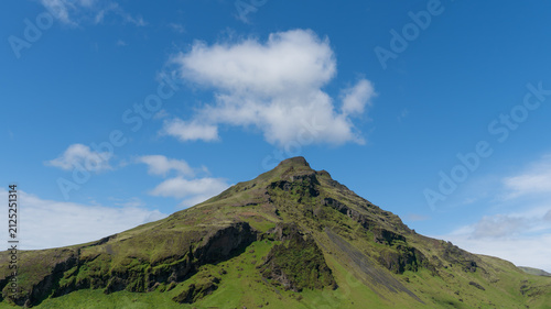 Bild auf Leinwand Ein grün bewachsener Vulkan vor blauem Himmel mit wenigen Wolken in Island