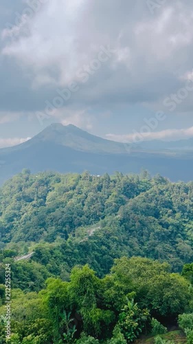 Panoramic aerial view of a lush green mountain valley covered in tropical forest, with a caldera in the distance and dramatic clouds rolling over the peaks, Bali highland landscape