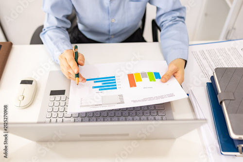 Woman analyzes charts on a laptop. She studies data with focused intent. Desk holds plants, notes, and a phone. Professional workspace with soft lighting

