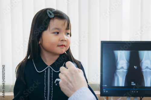 A kind, elderly physician gently comforts a young girl during a medical examination in his office, while her mother stands close by offering support, with an X-ray scan displayed on the computer scree