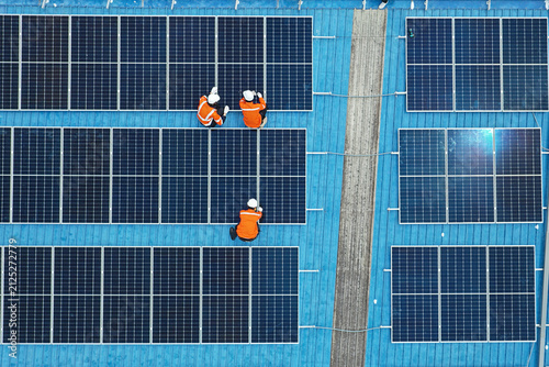 Aerial view of technicians installing solar panels on blue roof. Engineers maintaining solar energy system on a building rooftop. Top down view of workers in orange uniform fixing solar panels. 