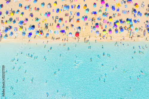 Aerial drone view of people relaxing on a white sand beach under colorful umbrellas. Bright sunny summer vacation scene by turquoise crystal clear sea water.