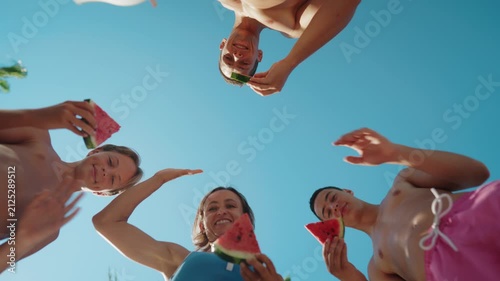 Low-angle view of a family standing in a circle outdoors on a sunny summer day, holding fresh watermelon slices and enjoying a healthy snack together. The camera looks up toward the blue sky, creating